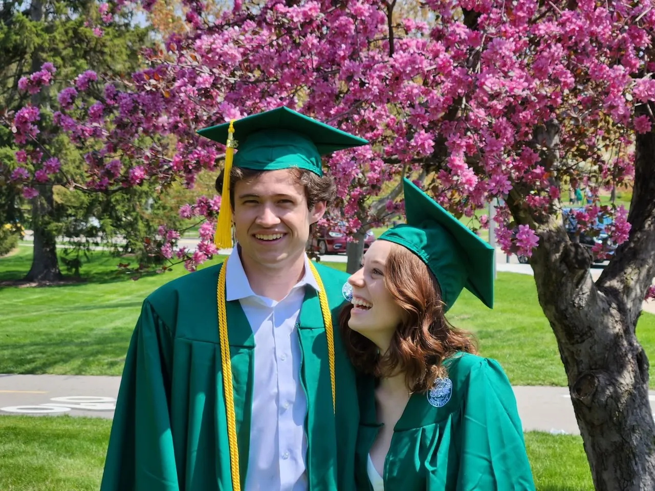 Jess looking up at Josh, both smiling in their MSU graduation caps and gowns in front of a pink flowering tree.