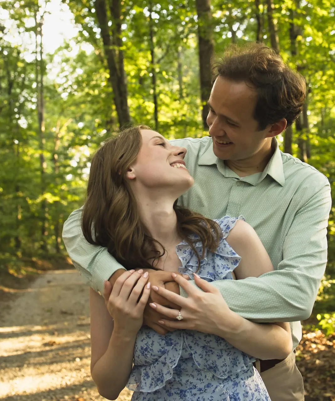 Josh holding Jess from behind in a wooded area, both smiling at each other